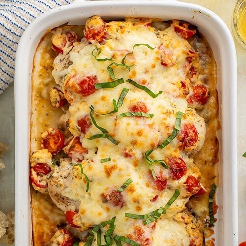 Overhead image of chicken breasts in a white baking dish topped with cherry tomatoes, shredded basil and melted cheese.