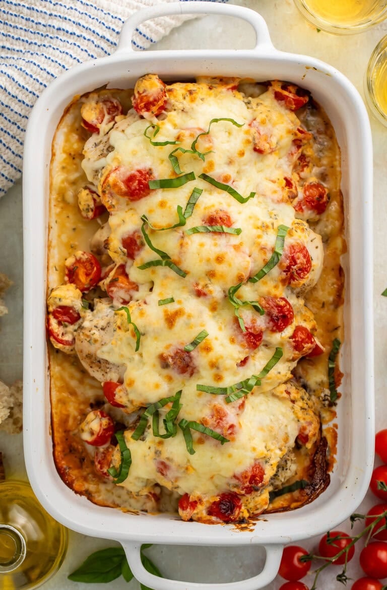 Overhead image of chicken breasts in a white baking dish topped with cherry tomatoes, shredded basil and melted cheese.