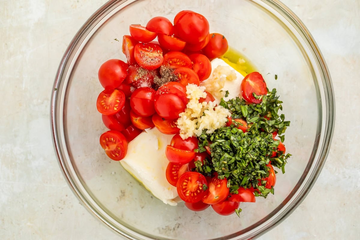 Halved cherry tomatoes, cream cheese, shredded basil and minced garlic in a glass bowl.