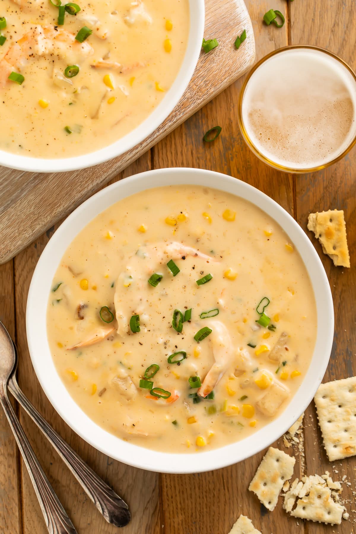 Bowls of corn and shrimp chowder topped with sliced green onion. Crumbled crackers and a glass of beer are off to one side.