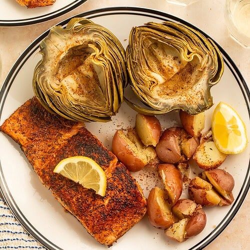 Plate of steamed artichokes, red potatoes and spiced salmon served with lemon wedges and butter.