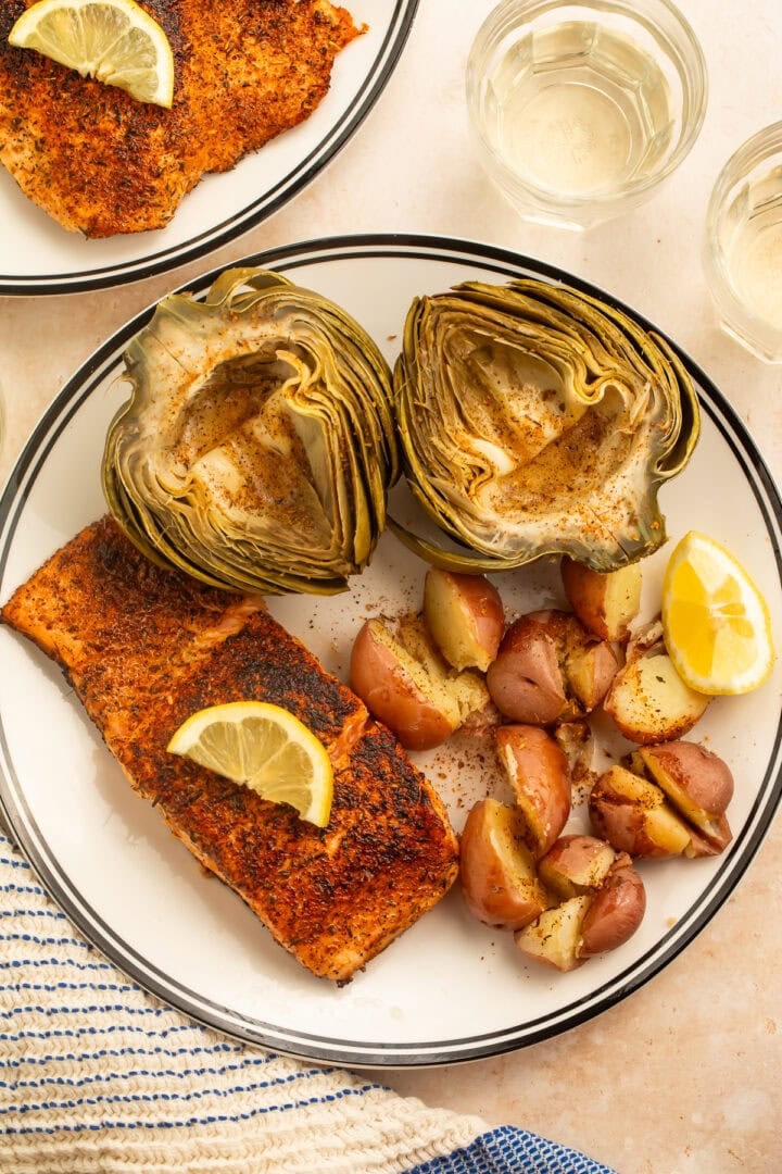 Plate of steamed artichokes, red potatoes and spiced salmon served with lemon wedges and butter.