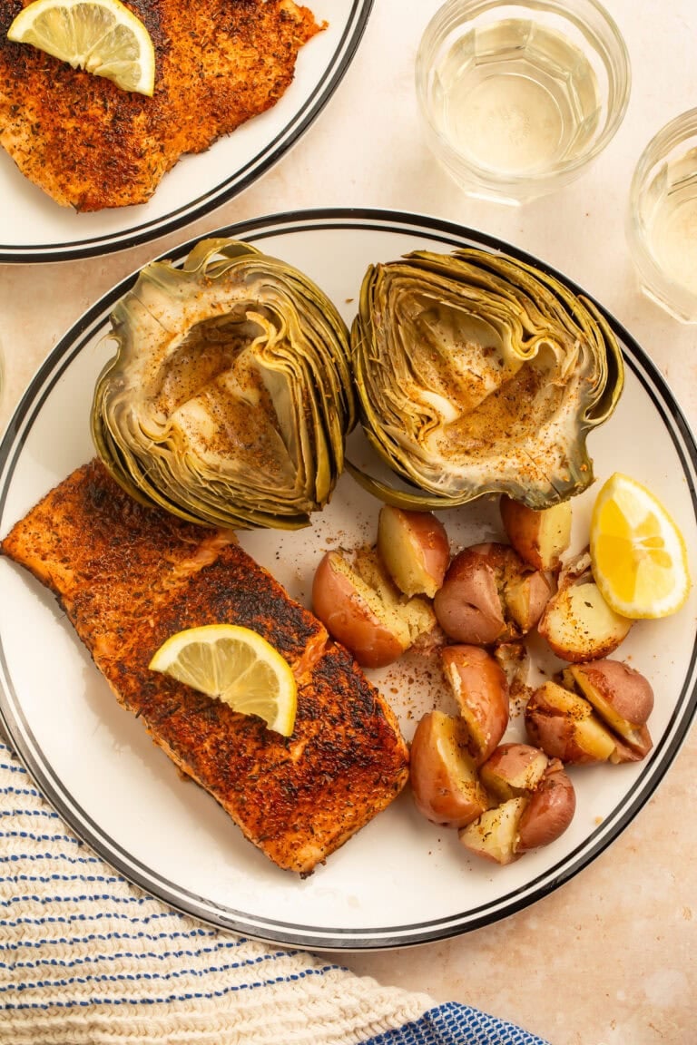 Plate of steamed artichokes, red potatoes and spiced salmon served with lemon wedges and butter.