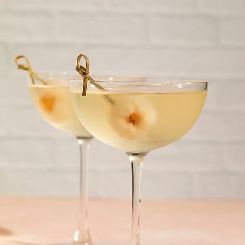 Side-on view of a pair of lychee martinis against a white brick background.