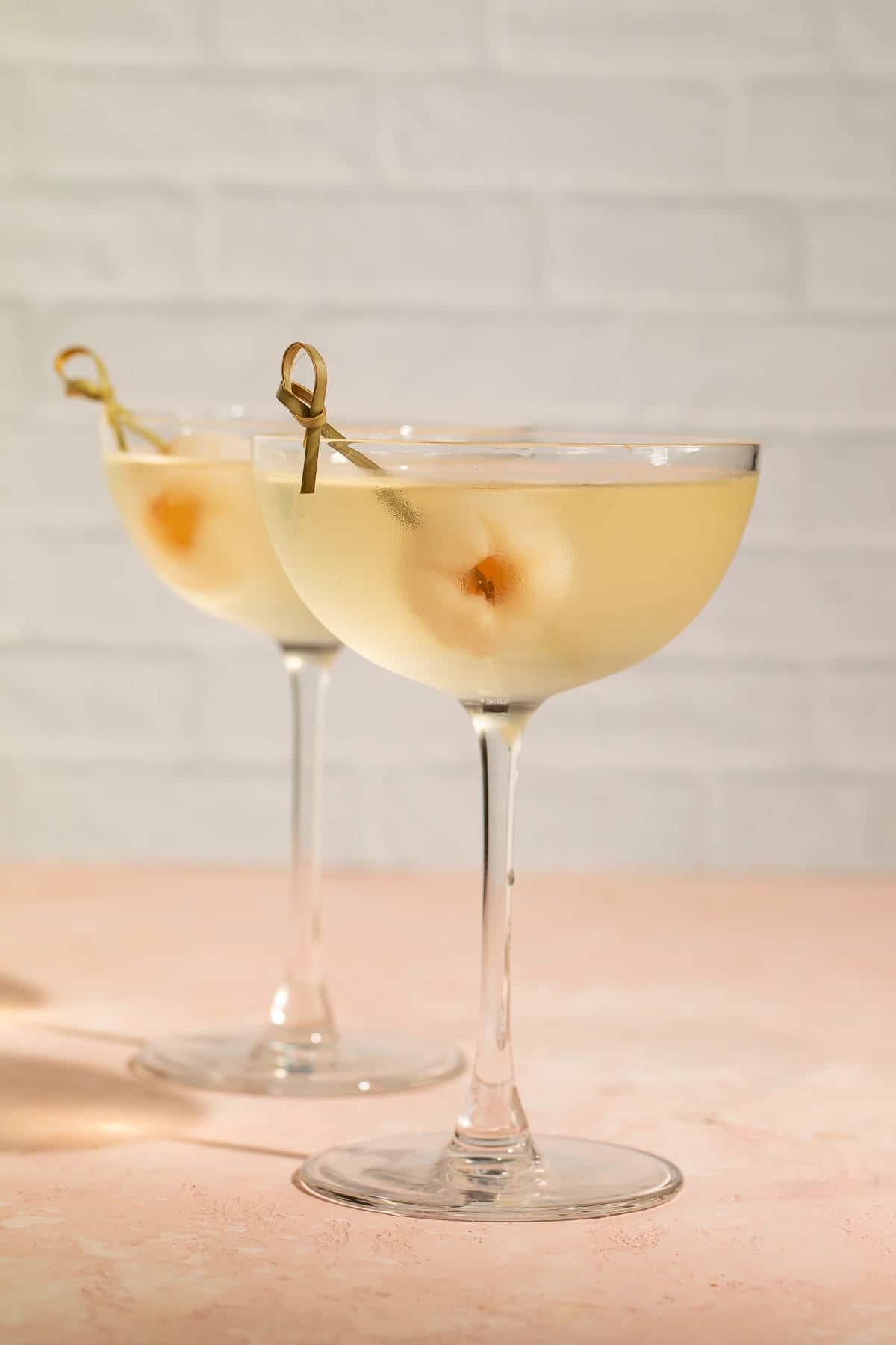 Side-on view of a pair of lychee martinis against a white brick background.