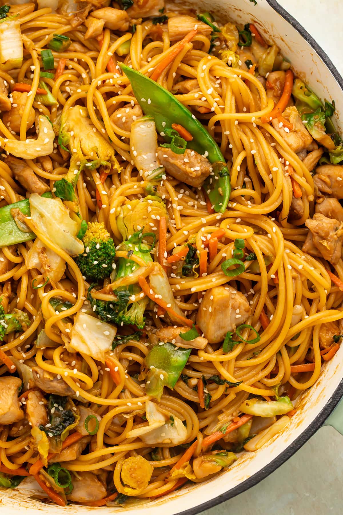 Close up of a chicken and vegetable lo mein in a skillet topped with scattered sesame seeds.