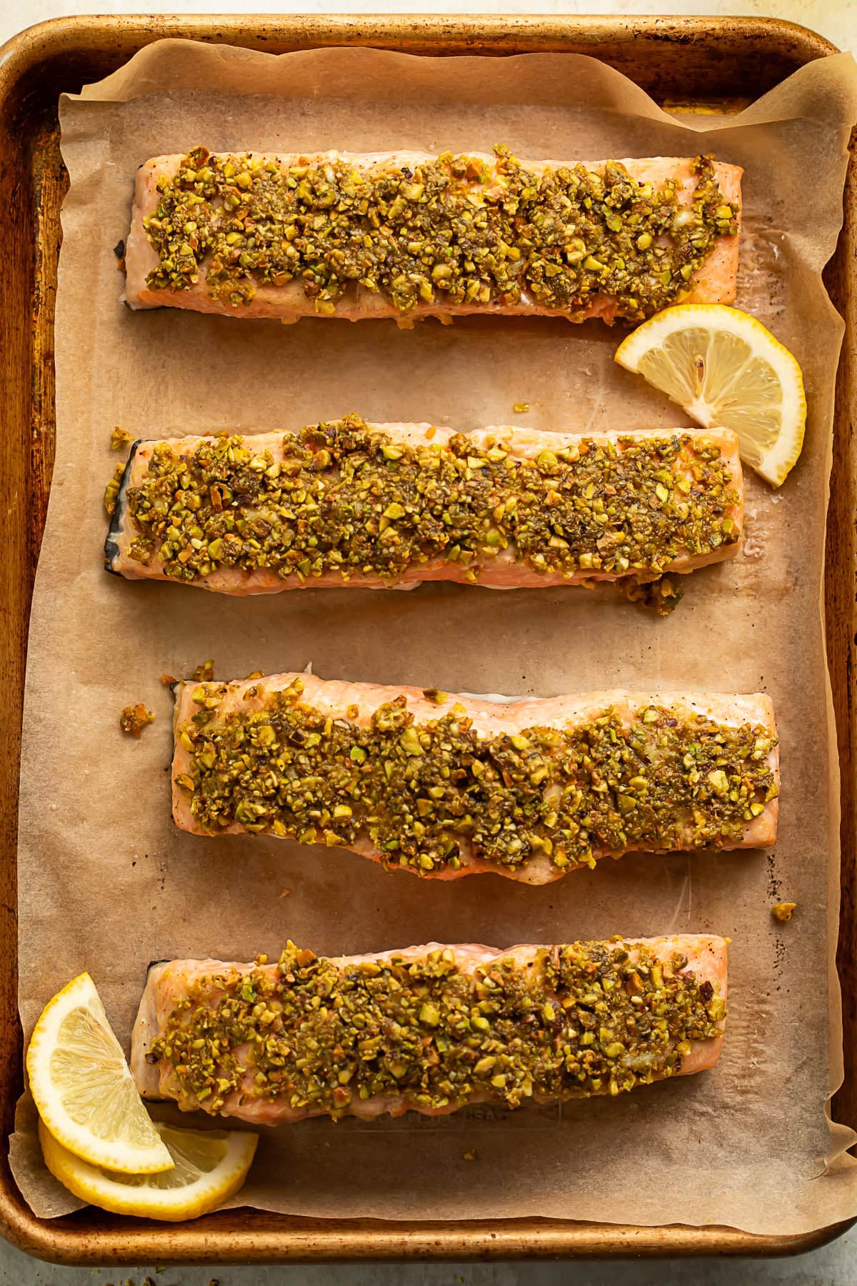 Parchment lined sheet pan with four salmon fillets with a pistachio crust. The tray is garnished with lemon slices.