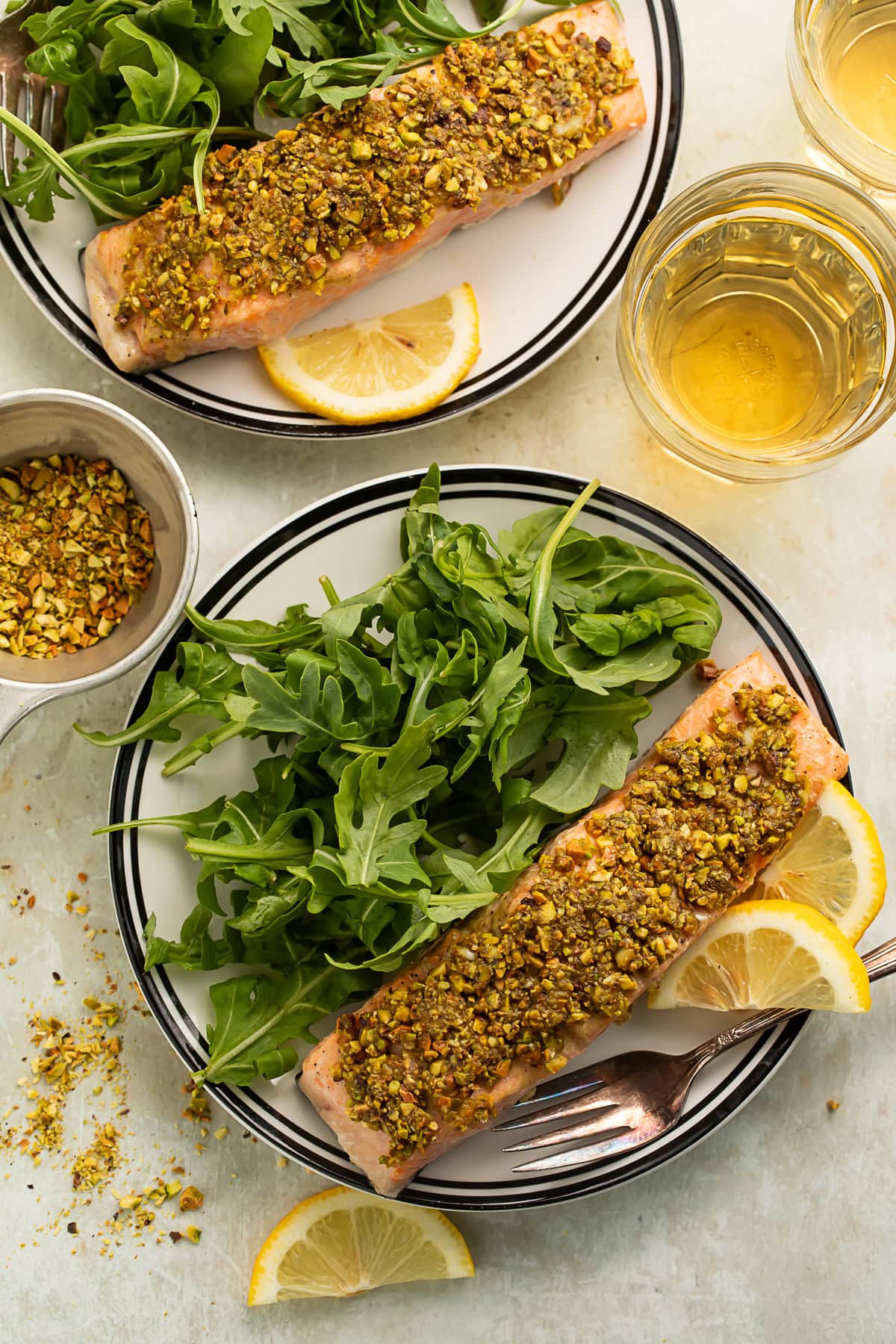 Two plates of salmon with a pistachio crust, tangled rocket and lemon slices. There is a measuring cup of crushed pistachios off to one side as well as another plate of salmon and glasses of white wine.