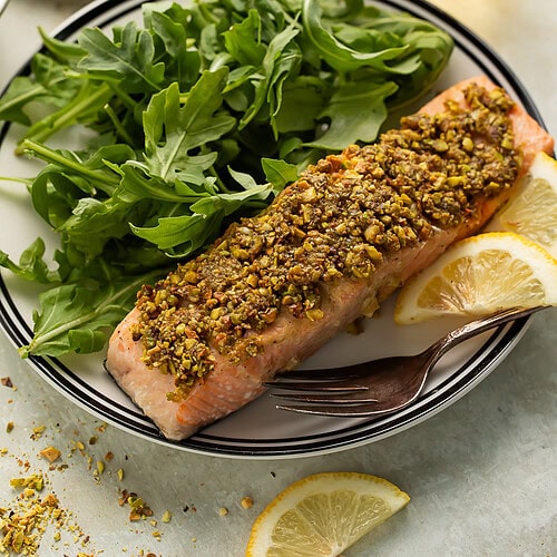 Pistachio crusted salmon on a plate with arugula and lemon slices. There is another plate of salmon and salad and a tumbler of white wine in the background.