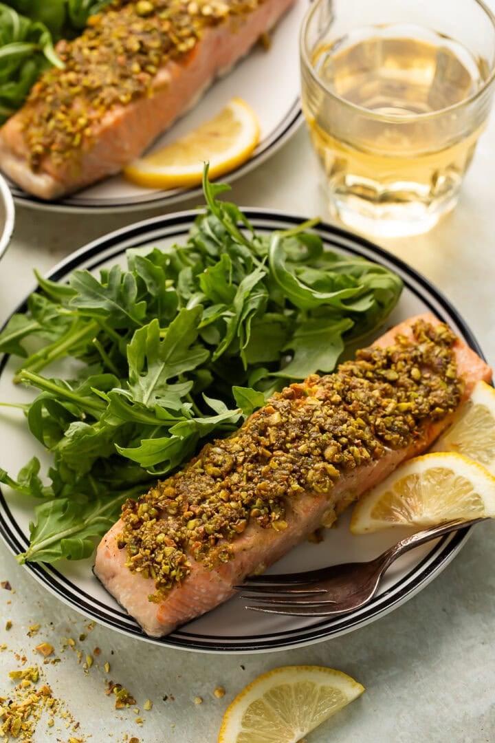 Pistachio crusted salmon on a plate with arugula and lemon slices. There is another plate of salmon and salad and a tumbler of white wine in the background.