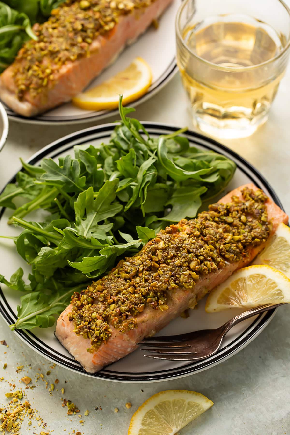 Pistachio crusted salmon on a plate with arugula and lemon slices. There is another plate of salmon and salad and a tumbler of white wine in the background.