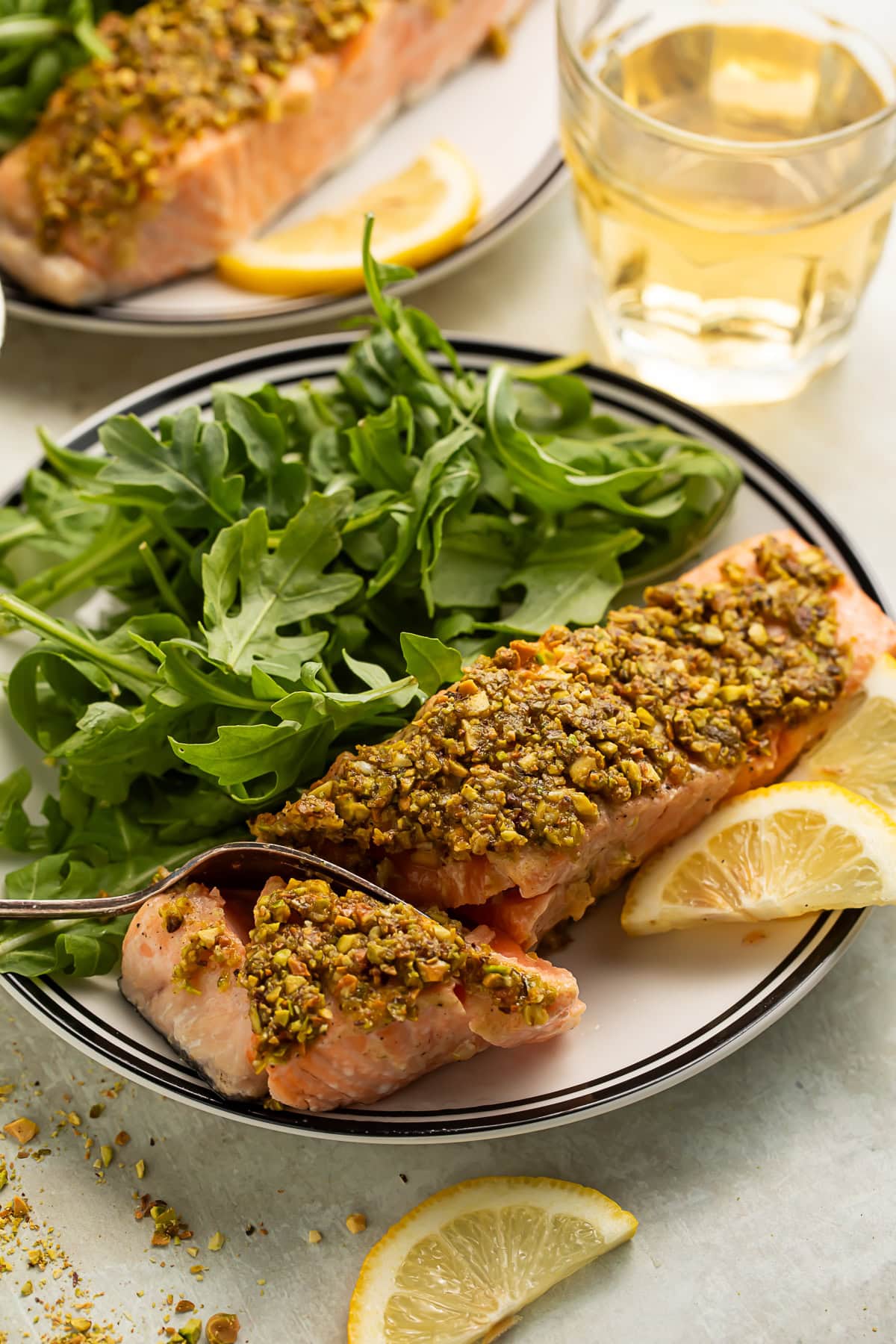 Side-on view of a fork taking a bite out of a fillet of salmon with a pistachio crust. On the plate is arugula and lemon slices.