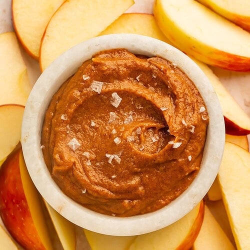 Raw salted caramel dip in a white bowl topped with flaky sea salt, surrounded by sliced apples