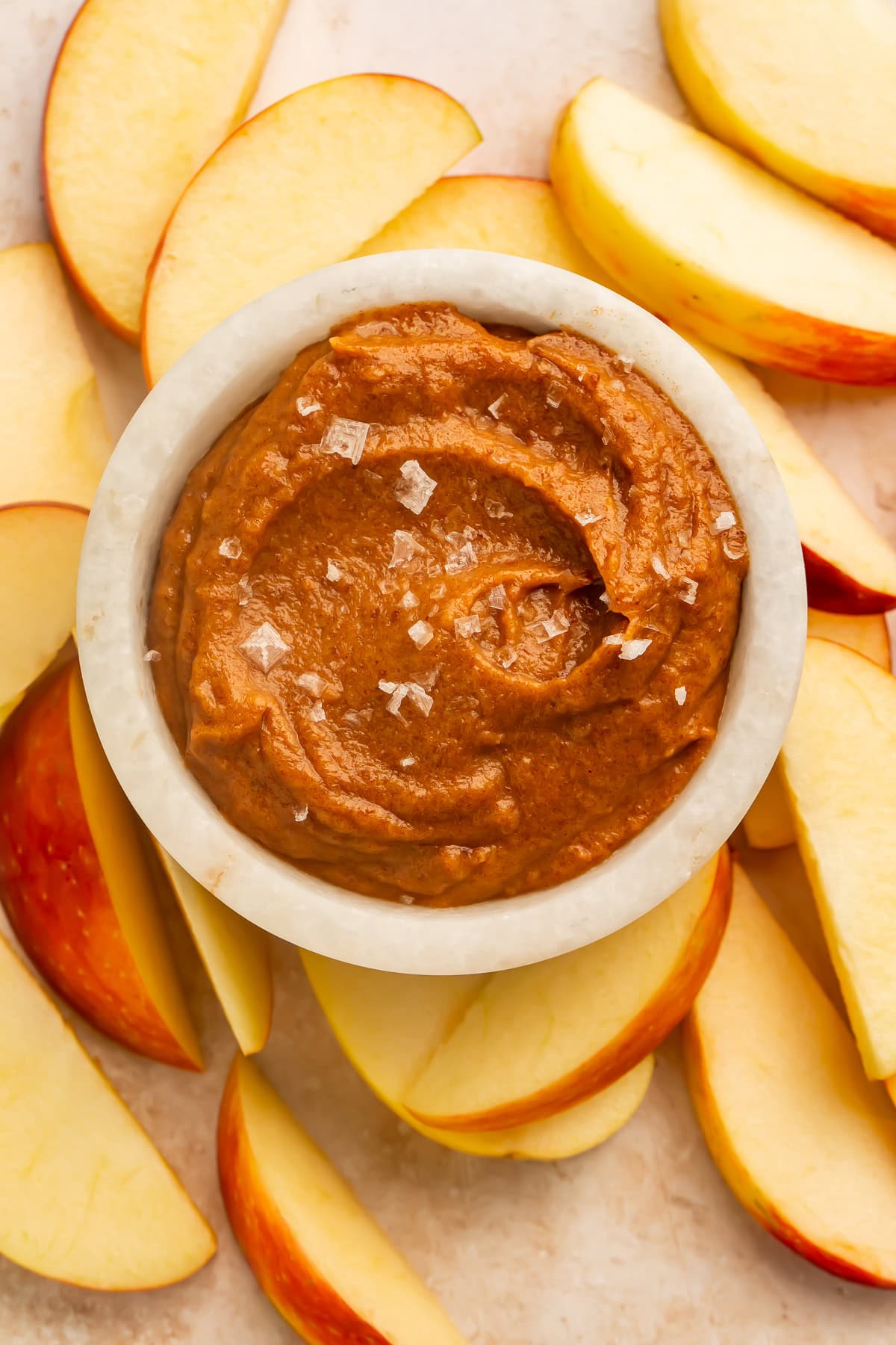 Raw salted caramel dip in a white bowl topped with flaky sea salt, surrounded by sliced apples