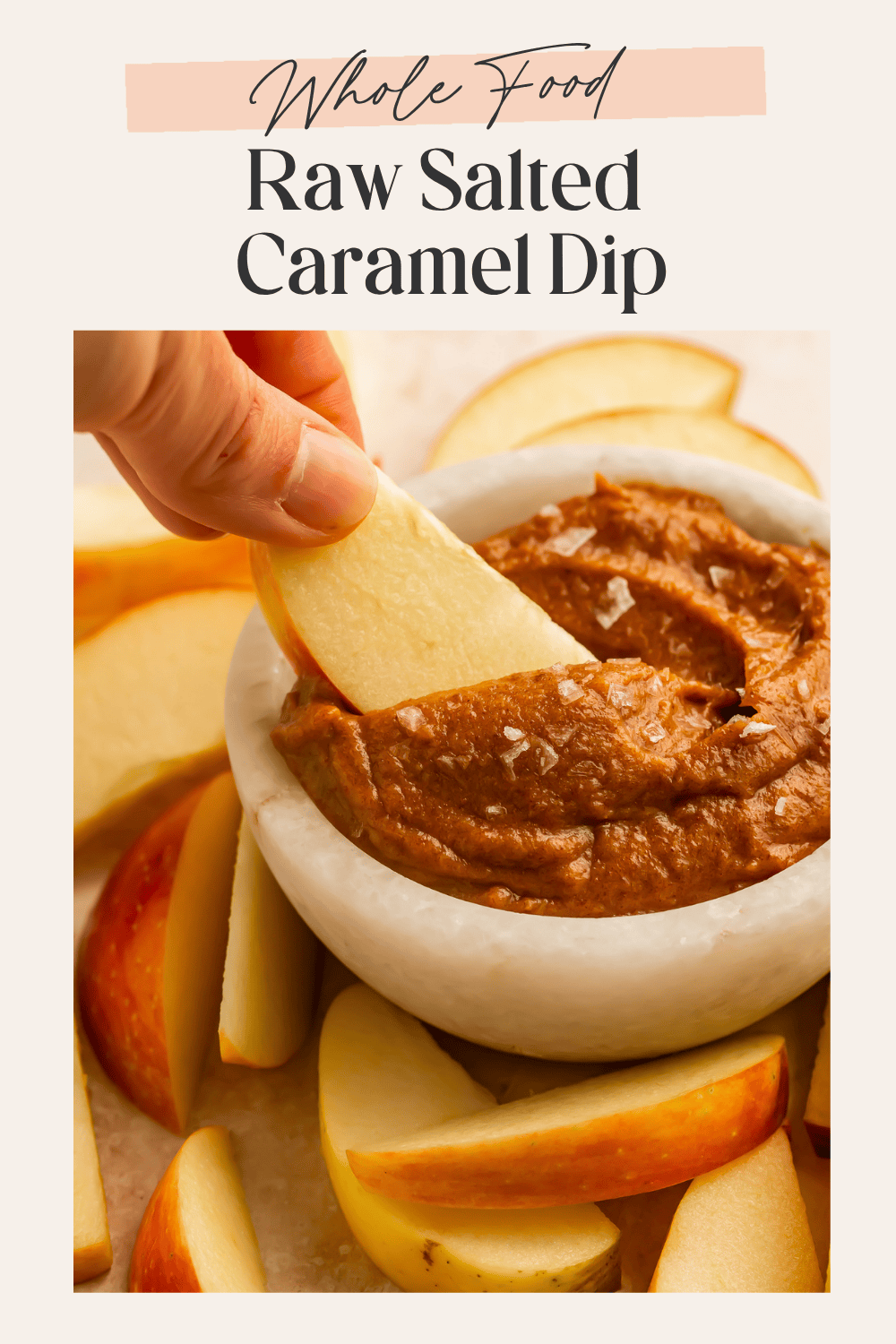 Hand dipping an apple slice into a white marble bowl of raw salted caramel dip surrounded by apple slices