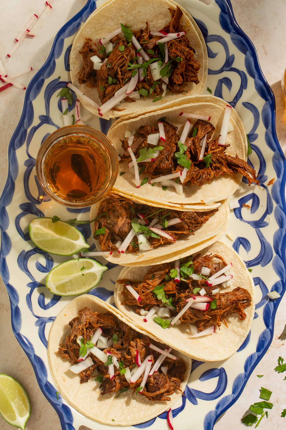 Overhead image of a blue and white plate of crockpot beef barbacoa tacos served with chopped radishes and cilantro, and lime wedges.