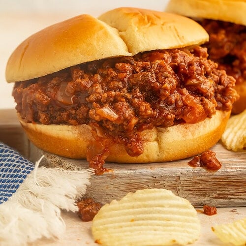 Side-on close up sloppy joe on a wooden board with ruffled potato chips in the foreground.