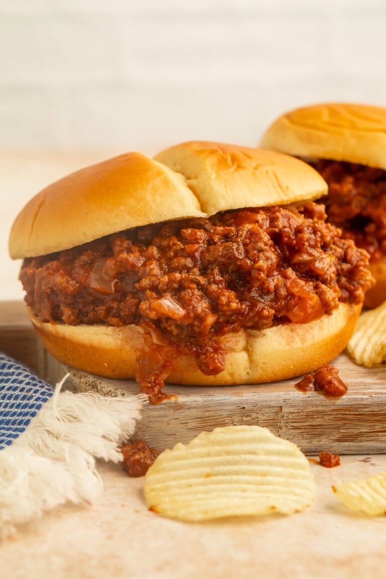 Side-on close up sloppy joe on a wooden board with ruffled potato chips in the foreground.