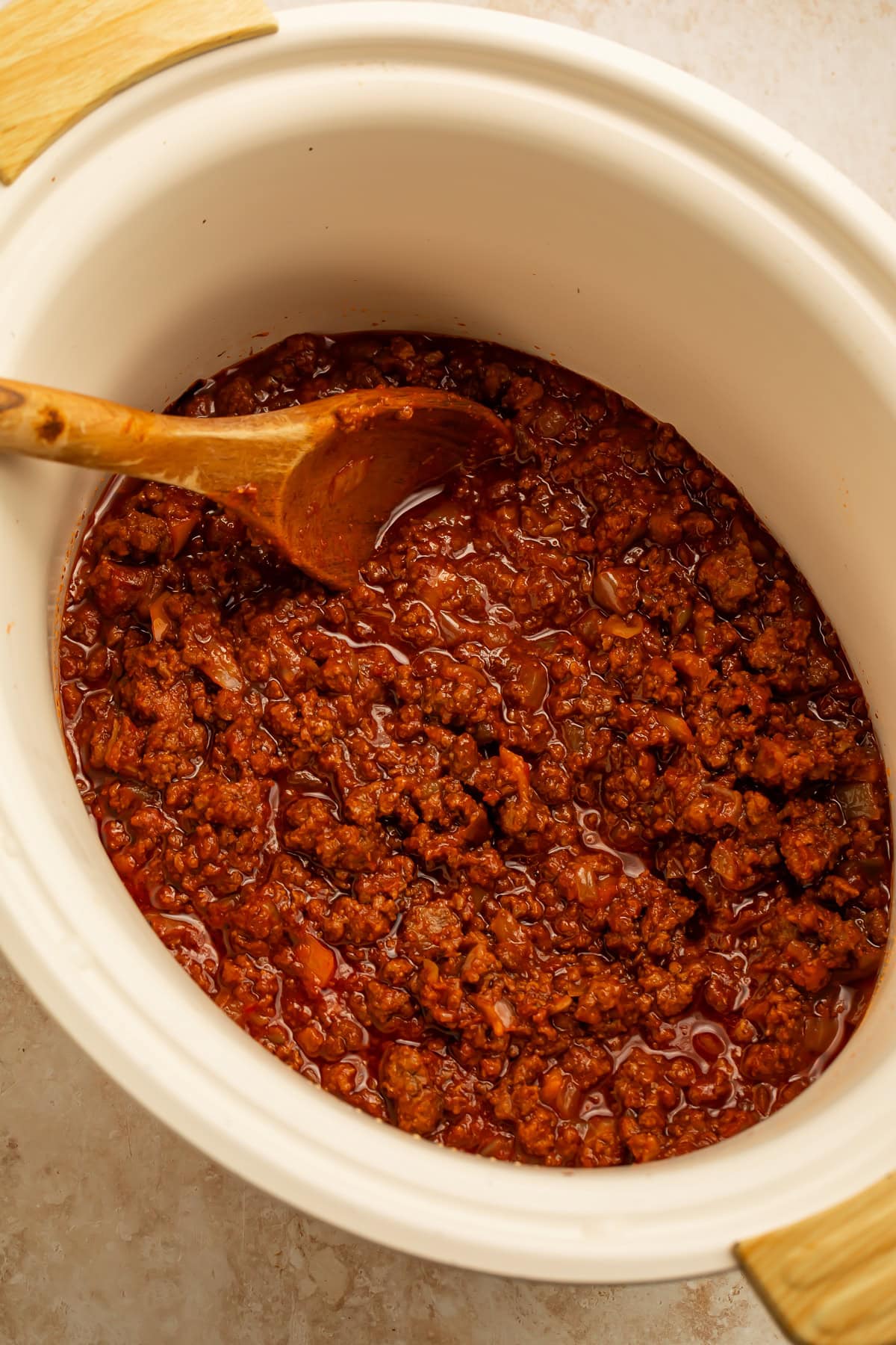 Close-up of sloppy joe filling in a white Crockpot insert with a wooden spoon.