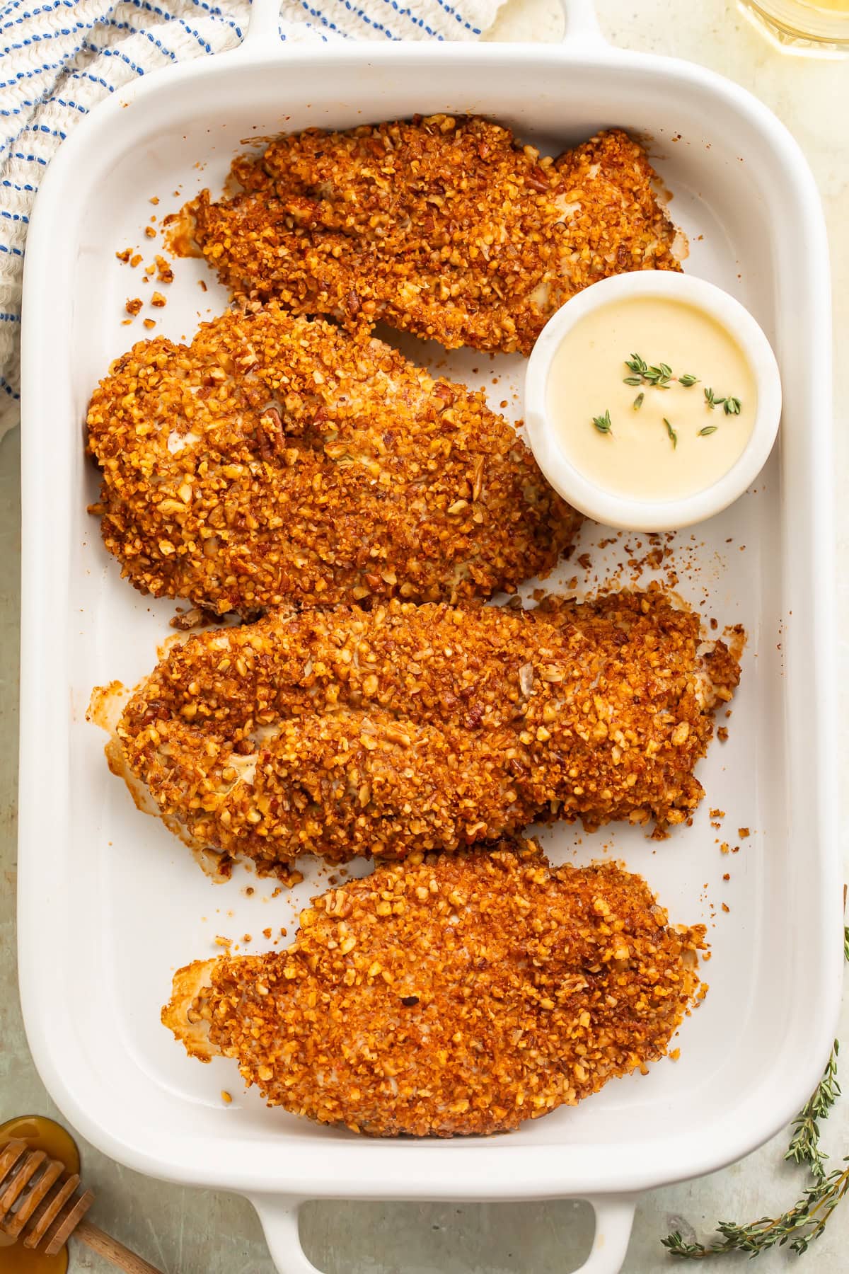 White baking dish of pecan chicken breasts with a small pot of creamy honey mustard sauce on one side.