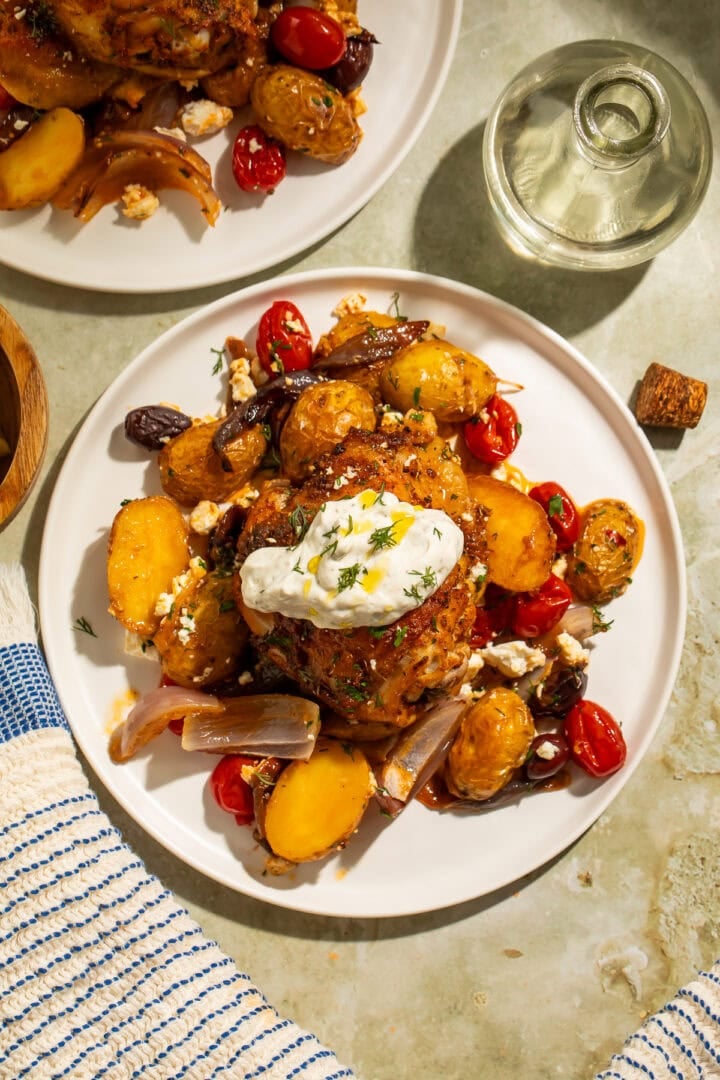 Greek seasoned chicken thigh on a white plate with roasted veggies and feta, topped with a dollop of tzatziki.