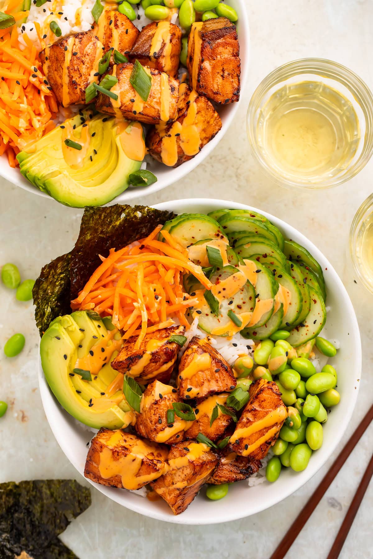 Overhead image of two bowls of spicy salmon bites over rice with nori, carrot, cucumber, avocado and edamame.