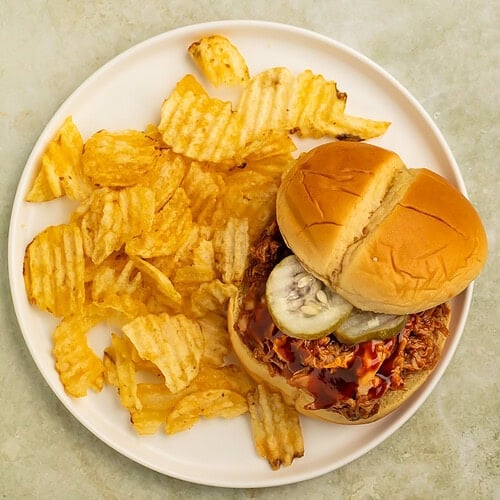 Smoky BBQ pulled pork sandwich with pickles served alongside ridged potato chips.
