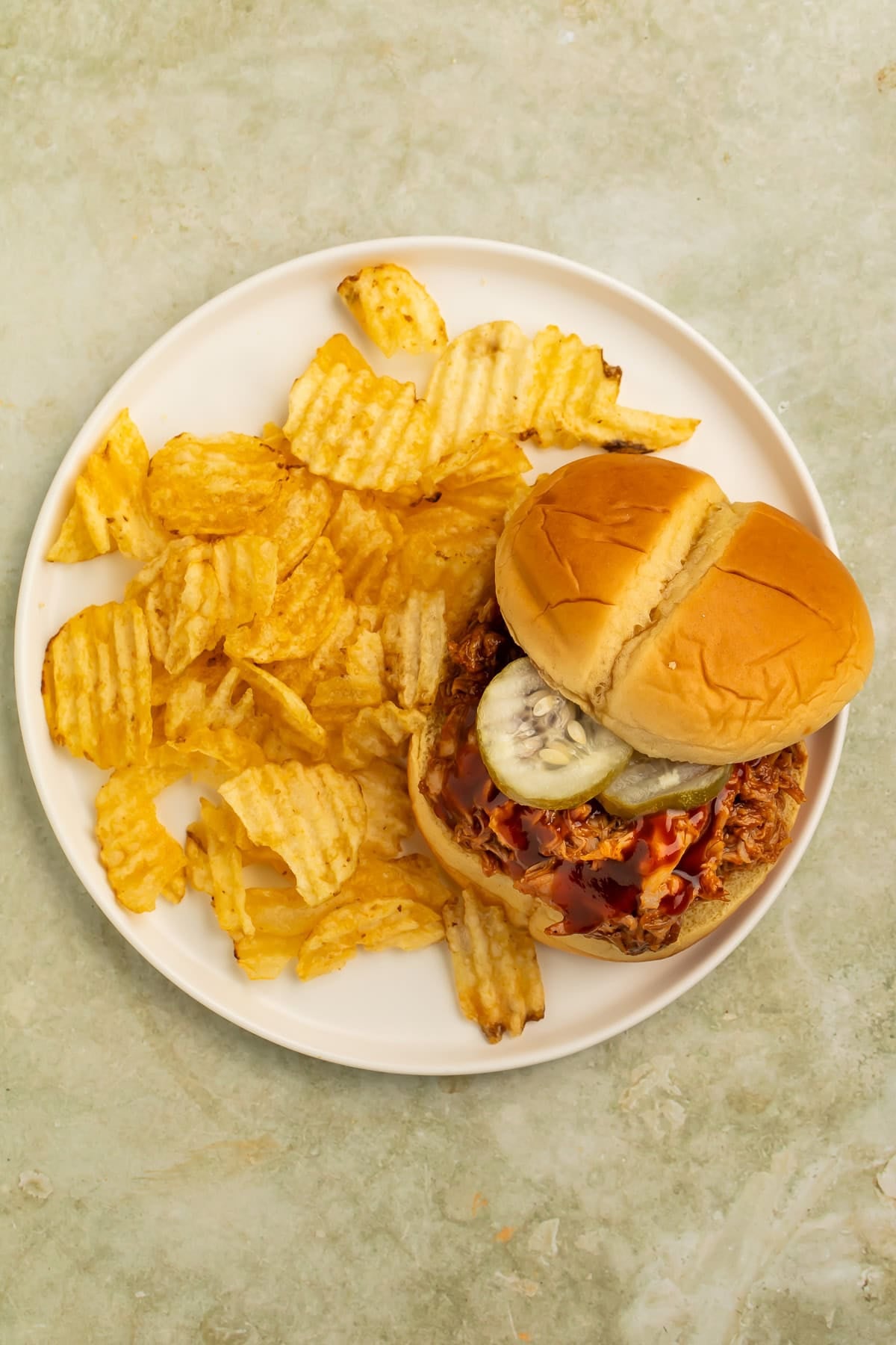 Smoky BBQ pulled pork sandwich with pickles served alongside ridged potato chips.