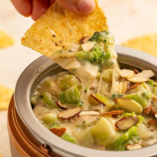 Hand with a tortilla chip scooping broccoli dip out of a mini Crockpot.