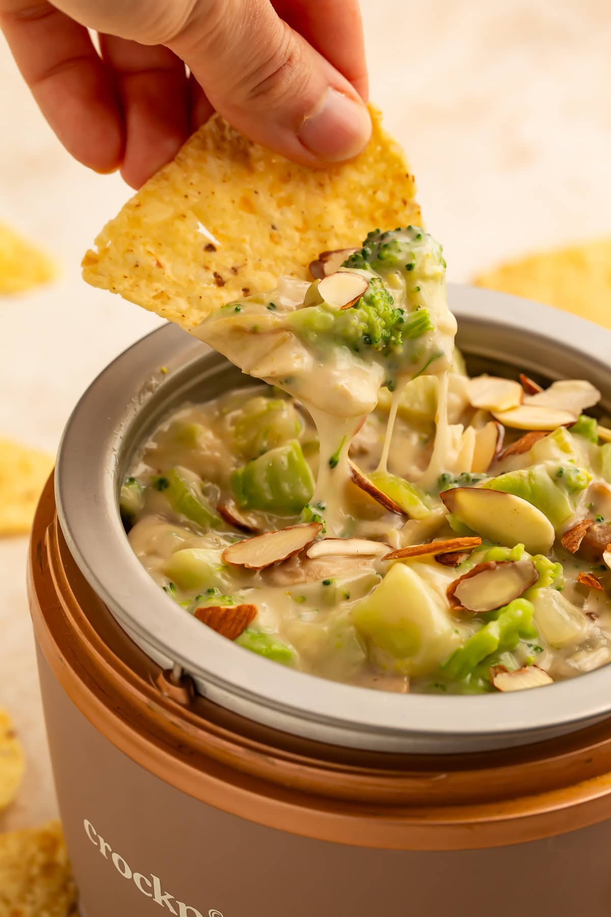 Hand with a tortilla chip scooping broccoli dip out of a mini Crockpot.