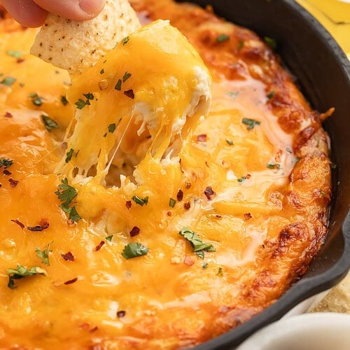 A hand scooping up hot honey chicken dip from a cast iron skillet with a tortilla chip.