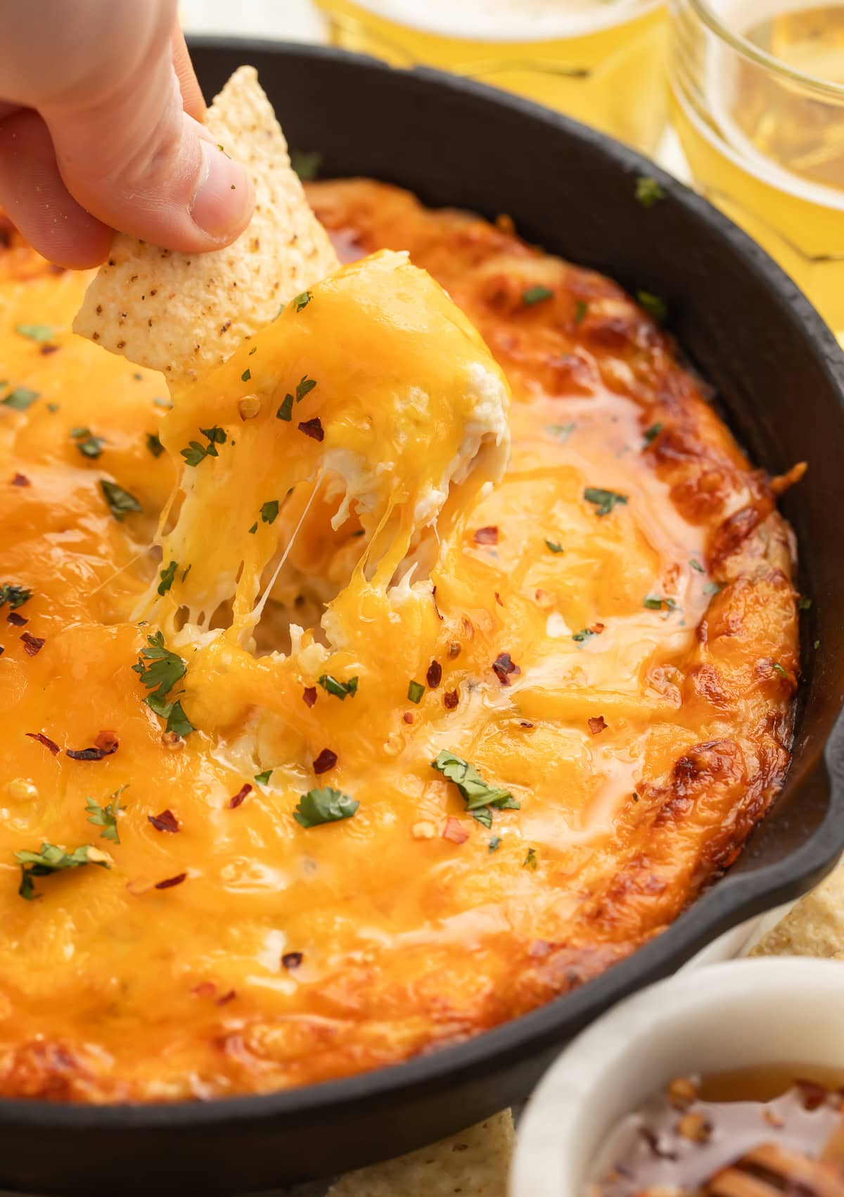 A hand scooping up hot honey chicken dip from a cast iron skillet with a tortilla chip.