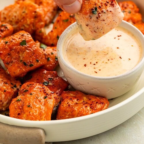 A hand dipping a Cajun honey-glazed salmon bite in a pot of creamy sauce.