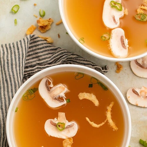 Two bowls of clear soup with sliced mushrooms, sliced green onions and crispy onions.