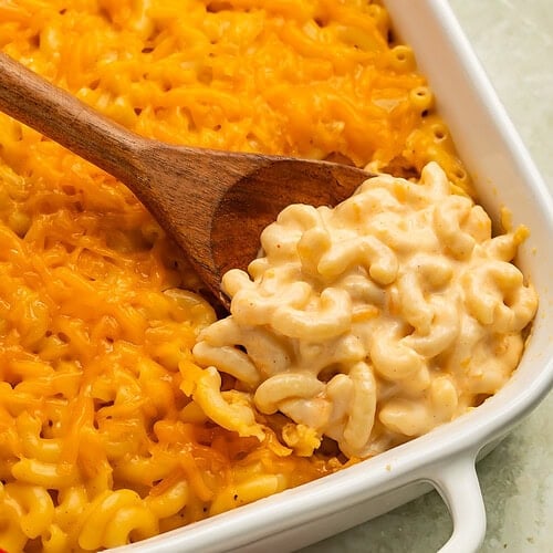 Wooden spoon scooping macaroni and cheese out of the white baking dish.