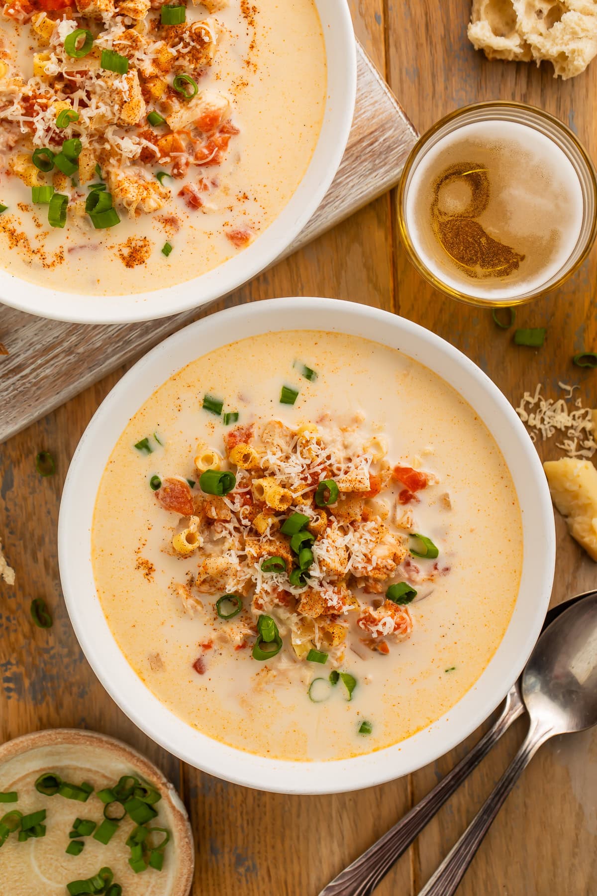 Two bowls on a wooden table of creamy chicken and tomato soup with Cajun seasoning. There is a glass of beer and a torn baguette on the table.