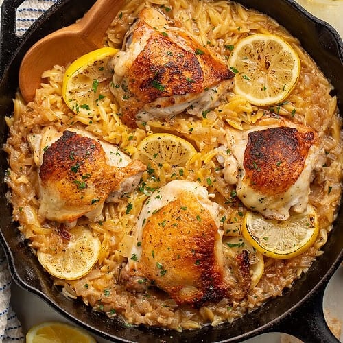 Case iron skillet of chicken thighs, lemon and orzo surrounded by glasses of wine, lemon wedges and crusty bread.