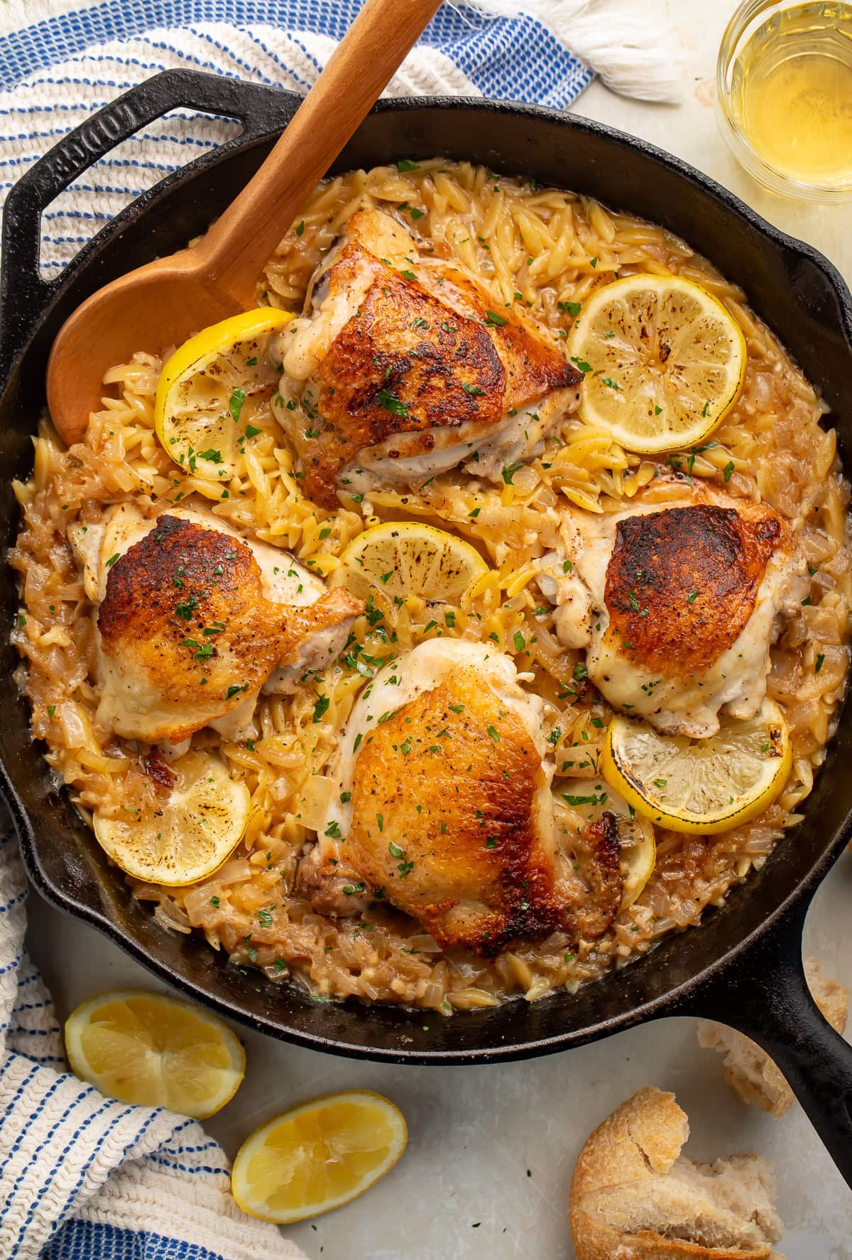 Case iron skillet of chicken thighs, lemon and orzo surrounded by glasses of wine, lemon wedges and crusty bread.