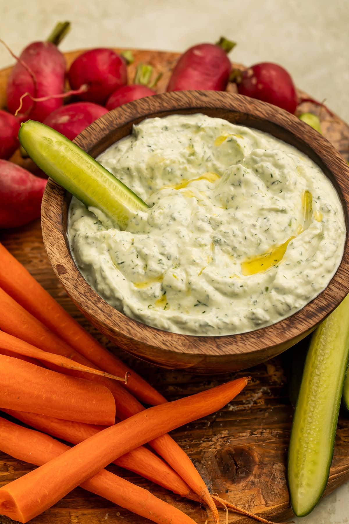 Cucumber spear dipped into a creamy cottage cheese dip in a wooden bowl, surrounded by crudites.