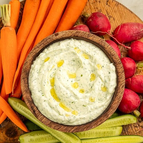 Bowl of creamy cottage cheese herb dip in a wooden bowl on a board with cucumber, carrots and radishes.