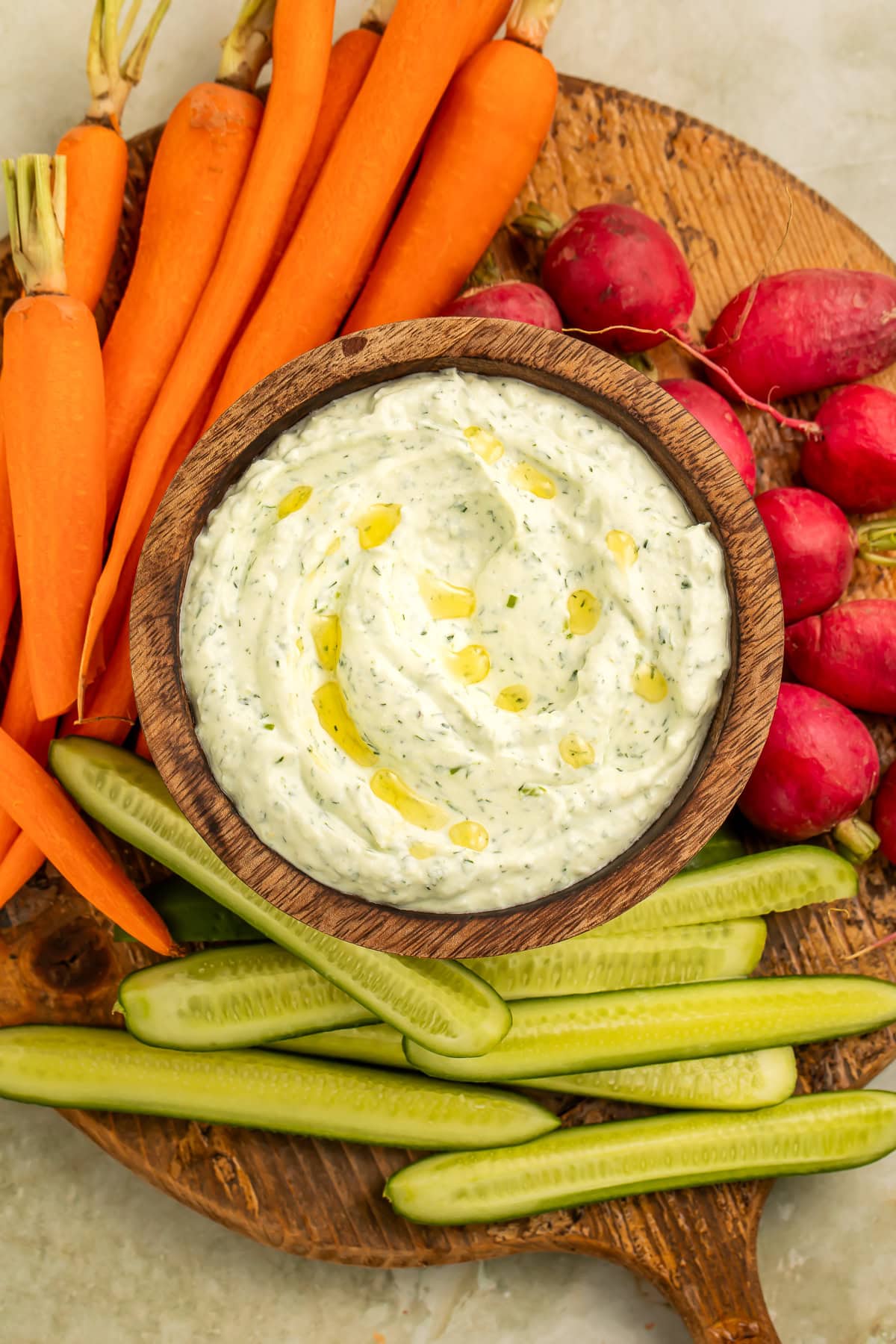 Bowl of creamy cottage cheese herb dip in a wooden bowl on a board with cucumber, carrots and radishes.