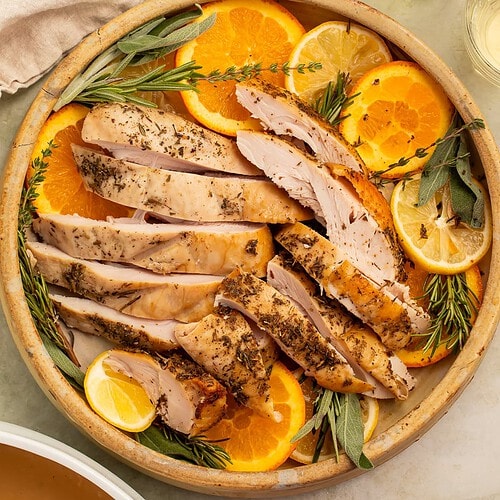 Sliced turkey breast on a serving platter with sliced oranges and woody herbs. There is a white boat of gravy on the side.