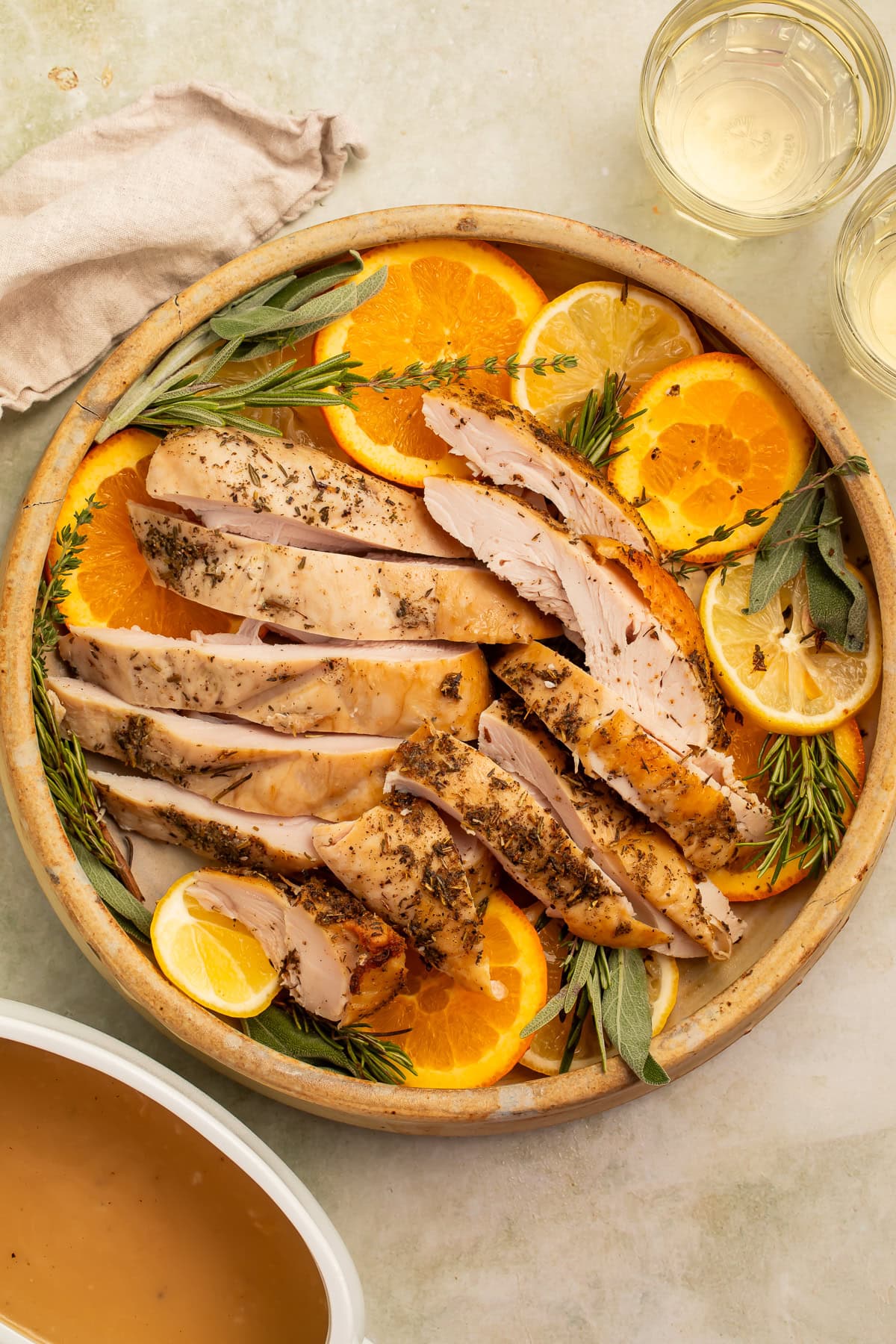 Sliced turkey breast on a serving platter with sliced oranges and woody herbs. There is a white boat of gravy on the side.