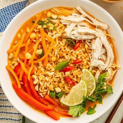 Close up of a bowl of peanut butter ramen topped with shredded carrots, bell peppers, lime wedges and shredded chicken.